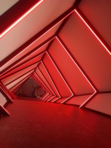 Intérieur d'un couloir aux murs géométriques, éclairé par des lumières rouges.